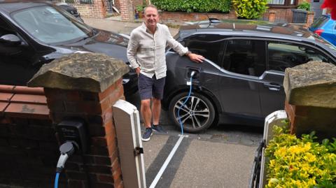 Chris stands on the pavement next to his black electric car, he wears navy shorts and a beige long sleeved shirt. The car is parked on the street, and the blue charging cable runs into a gully through the pavement, covered by a narrow metal strip. It connects to the wall of a terraced house which has red brick walls.