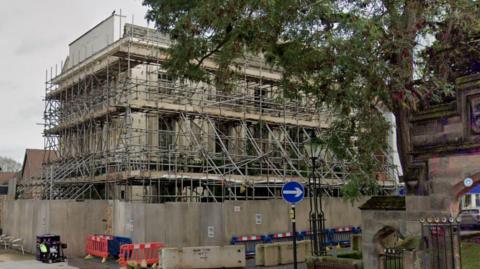 The hotel which is surrounded by a fence and covered in scaffolding. A tree can be seen hanging over head and there is also a road sign in view. 