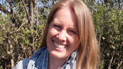A woman with blonde hair, wearing a white top and a scarf, smiles at the camera. There are trees and shrubbery behind her.