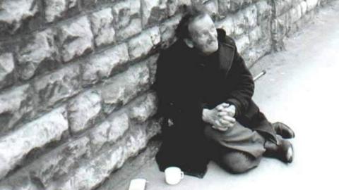 A black and white photo of Bill Clements sitting on the pavement next to a wall. He is wearing a long black coat, dark trousers and leather shoes. His hands are clasped together and there is a teacup on the floor next to him.