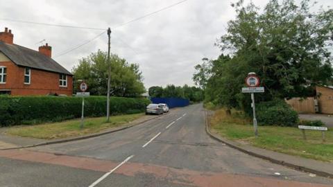 Streetview image of the entrance to Baines Lane, a semi rural road with hedges and trees 