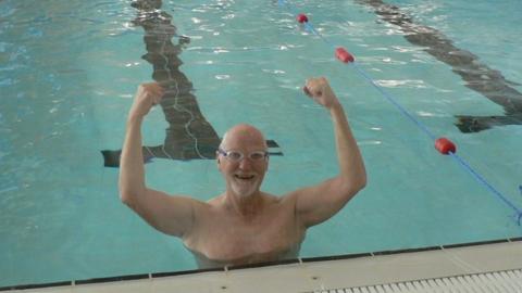 Bob Young at the shallow end of the pool with his arms in the air, smiling and wearing goggles.
