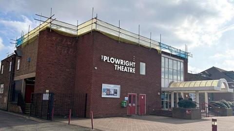 The exterior of the Plowright Theatre in Scunthorpe. The large red -rick building has large white lettering on the front along with two red-coloured doors. The right of the building has a large glass canopy leading to the main doors and  floor-to-ceiling glass windows. Scaffolding, covered with blue and yellow mesh, is visible on the roof.