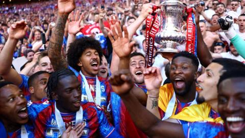 Crystal Palace FC players gather together on the pitch in front of crowds, all smiling, one holding the FA Cup trophy in the air 