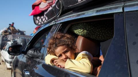 A young girl, wearing a yellow jumper leans her cheek on her arms crossed in the open window of a car that is piled with belongings, other vehicles visible ahead of it, as Gazans wait to return to the north of the territory