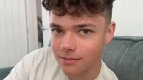 A teenage boy with brown curly hair, short on the sides. He is wearing a white Under Armour t-shirt and a thin chain as he sits on an armchair.