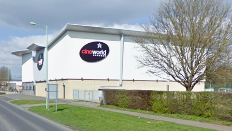 A Google Streetview image of the outside of Cineworld, take from the middle of the road. It is a large white and cream coloured building with the black, red and white Cineworld Cinemas logo on front and side of it. There is a tree and a row of bushes on the right side of the image.
