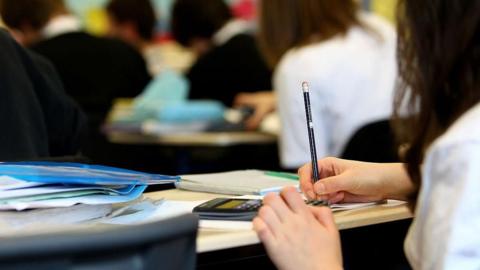 Pupils in a classroom. In the foreground is the side of a pupil holding a pencil. There is a calculator, books and papers on the desk. 