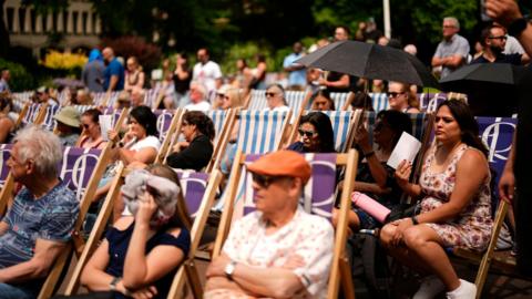 People sit in deckchairs during a hot, sunny day at the Victoria Embankment with some shielding themselves using umbrellas, paper fans, or clothing to cope with the heat.