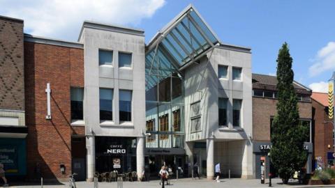 An entrance to Chantry Place shopping centre. The facade is red brick and grey render with a glass roof. Caffe Nero is located at the front of the centre and three people are waling by