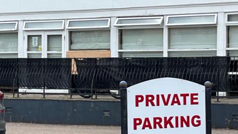 a private parking sign sits in front of a fence which has a set of windows behind it, one of which has been removed after being broken
