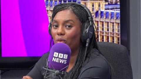 A black woman with long black hair sitting in a radio studio wearing headphones and talking into a microphone. She has black hair braids and is wearing a black top.