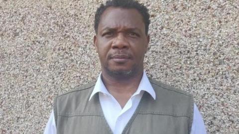 Charles Ehikioya seen standing in front of a textured beige wall. He is wearing a white collared shirt under a green vest with a small brown detail near the chest pocket.