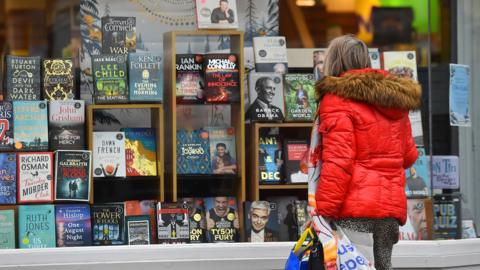 A person in a red puffer coat holds shopping bags as they look at book titles displayed in a window of a Waterstones branch