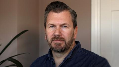 John Hammond head and shoulders picture looking at the camera with a neutral expression wearing a navy blue shirt with a white door, pink wall and plant behind him.
