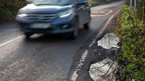 A car drives past two large potholes at the side of the road.