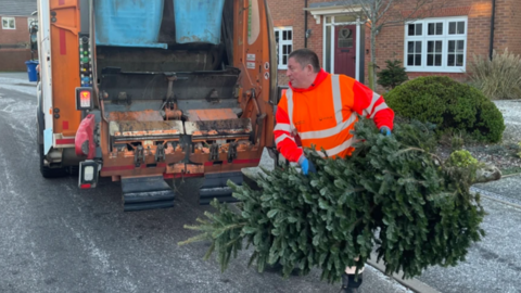 A man in a bright hi-viz jacket is lifting a large Christmas tree into the back of a wagon. The vehicle is parked outside houses