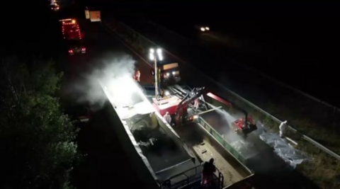 A night-time clean-up operation on a dual carriageway, viewed from an elevated position. A piece of heavy machinery with a lifting arm is positioned alongside the trailer and appears to be removing or handling material from inside it.
