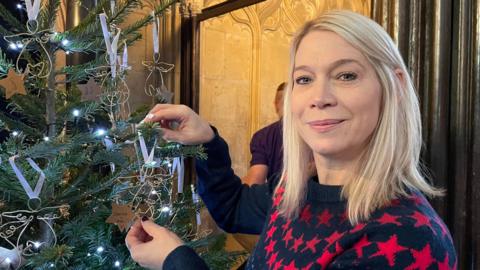 A fir Christmas tree on the left of the photo is decorated with angel figures made from sculpted golden wire. Each figure hangs on a white ribbon.  A cardboard star is pegged to the bottom with the name of a child written on it. A woman with blonde, shoulder-length hair is placing an angel on the tree and looking at the camera. She is wearing a dark blue jumper with red stars on it.