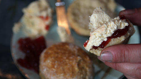 Part of a scone with jam and clotted cream on it (jam first, cream on top) held between two fingers. Out of focus in the back ground are more scones on a plate.