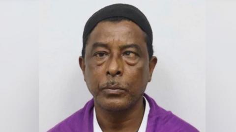 Headshot of a man looking straight ahead with a small black cap on his head, wearing a magenta shirt