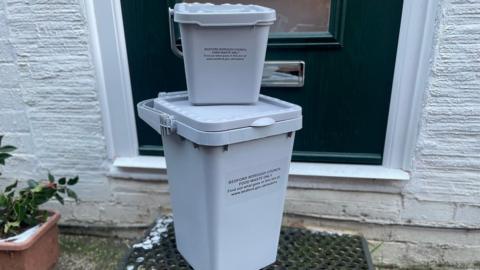 Two grey food caddie bins, outside a house, both stacked on top of each other. There is a front door behind them and on a dark mat. A plant is to the left. The house wall is painted white. 