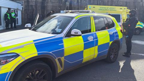 An officer is dressed in a black helmet with a black face covering which partially obscures the face and black clothing and is holding a black firearm and standing besides an emergency services vehicle. A Jersey Ambulance vehicle is behind the car. Two people with high-vis tops are in the background.