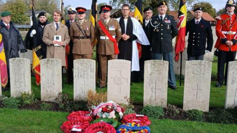 Seven identically shaped headstones in a row, facing forwards, engraved with a cross and words. Wreaths of poppies are laid in front of the newest grave, and behind the graves stand a group of people. One is a vicar wearing white robes and a black cassock, others are wearing brown or black miltary uniform and holding flags. One man appears to be a family member, standing holding an order of service.