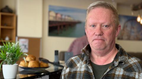A man in a plaid shirt standing in a cafe with a plate of scones in the background 