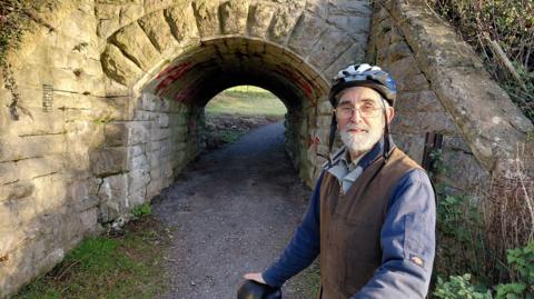 A man stands with a cycle helmet on, holding the saddle of a bike wearing a blue jacket and brown waistcoat. Next to him is a pathway with a tunnel leading to a green space