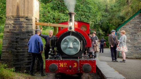 A small red steam locomotive stands in a countryside station next to a water tower. The locomotive has a face attached to the smokebox at the front, making it look like one of the characters from Thomas the Tank Engine. People in 1950s period costume stand on the platform