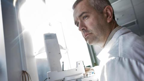 Dr Stephen Grimes, Associate Professor in Stable Isotope Geochemistry and Climate Change at the University of Plymouth. He is stood up and looking to the left. He is wearing a white lab coat. In front of him is a microscope and bright light. 