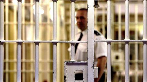 A prison officer walks through a prison with bars in the foreground.