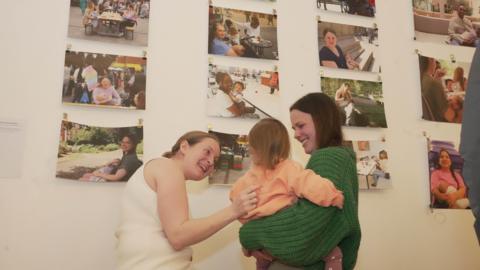 One woman leaning in towards a child and smiling. The child is being held by another woman with brown hair and a green jumper. Ona white wall in front of them are pictures of women breastfeeding in public