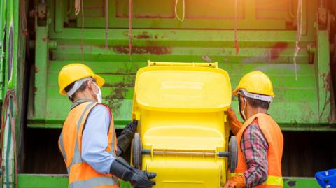 Two men, wearing yellow hard hats, holding a yellow bin as they tip it into an open green bin lorry 