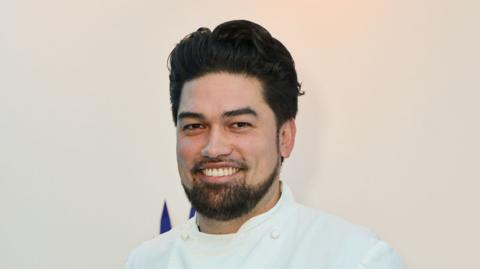 Luke Selby, wearing a chef's white outfit, smiles at the camera. He has dark hair and a short dark beard
