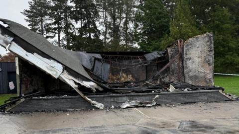 A burned out bandstand in Town Park in Harlow which is made from bricks and a metal structure. A fire has left the roof fully collapsed with burn marks all over it. 