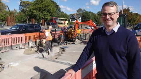 A construction site with several workers and machinery. In front of the works, a person wears black glasses and a blue jumper smiling at the camera. He is stood leaning on an orange barrier. Behind the barrier, three workers in high-vis vests are seen working working, with one using an orange digger. The background features a tree-lined street with a black car and a red double-decker bus.