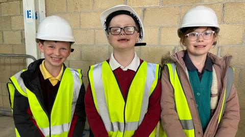 Three children smile at the camera - they are nine and ten years old. Two boys on girl. They are all wearing yellow high-vis vests and white hard hats. They stand in front of a wall.