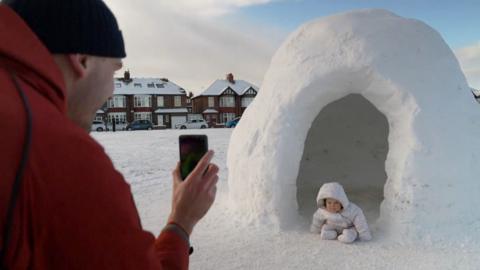 A man in a red winter coat takes a picture with his smartphone, of a baby sat in the entrance of an igloo.