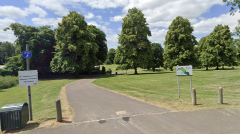 The entrance to Monkton Park as seen from an adjacent road. There is a winding tarmac path going into the park, which is broad and grassy, with neatly-space tall leafy trees in the background.