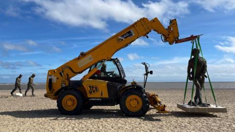 A yellow telescopic handler is parked on a beach.  Its fork is lifting a sculpture of a walking man which is standing on a platform.  Two walking men are visible behind the machinery.