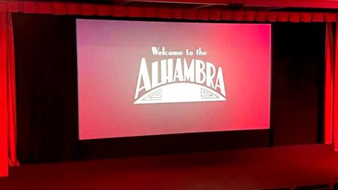 Cinema screen with Alhambra logo on it surrounded by red curtains