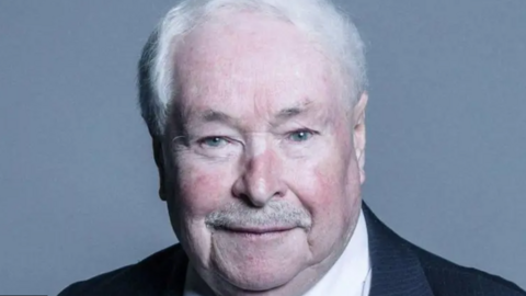 Wearing a dark suit and white shirt, Lord Doug Hoyle smiles as he looks straight ahead for the camera