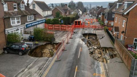 A massive sinkhole in the middle of a road, with houses to the left and right.