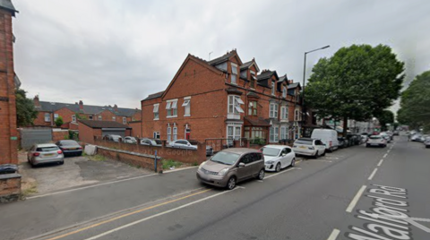 Row of red-brick terraced houses on a residential street with parked cars along the curb, a large leafy tree on the pavement, and overcast sky above.
