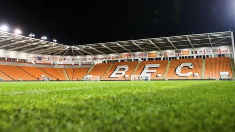 Floodlit Blackpool FC stadium with the pitch in the foreground with tangeirne seats and a white letters spelling out BFC.