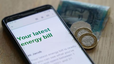 Energy bill on phone, with cash - a five pound note, two pound coins and fifty pence - on the side.