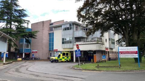 The exterior of Cheltenham General Hospital in Gloucestershire. It is a largely grey building with some cream panels, and there is one ambulance parked outside