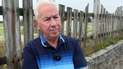 Norman Long standing on the roadside with his arms folded. He has white hair and is wearing a blue striped polo shirt